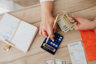 Mãos organizando dinheiro, recibos e uma calculadora sobre uma mesa de madeira.