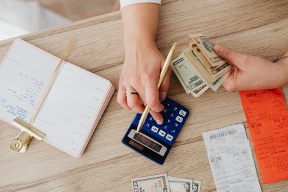 Mãos organizando dinheiro, recibos e uma calculadora sobre uma mesa de madeira.
