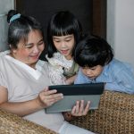 Adulta sentada com duas crianças, acompanhando o uso de um tablet em um ambiente doméstico.