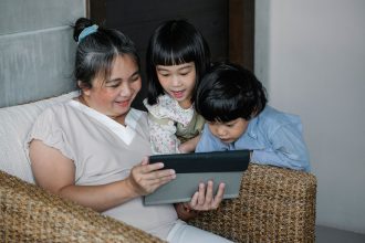 Adulta sentada com duas crianças, acompanhando o uso de um tablet em um ambiente doméstico.