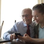 Homem idoso de óculos usa um smartphone enquanto recebe orientação da neta, sentados à mesa próxima a um laptop.