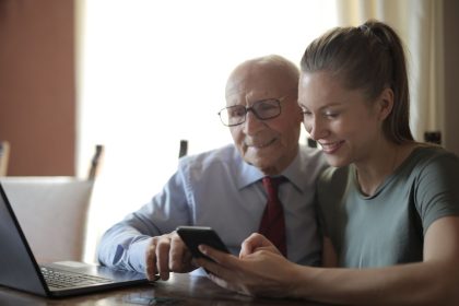 Homem idoso de óculos usa um smartphone enquanto recebe orientação da neta, sentados à mesa próxima a um laptop.