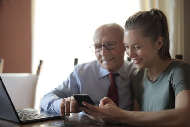 Homem idoso de óculos usa um smartphone enquanto recebe orientação da neta, sentados à mesa próxima a um laptop.